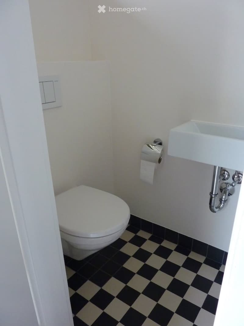 Small bathroom with white walls, black and beige checkered floor, toilet, sink, and toilet paper holder.