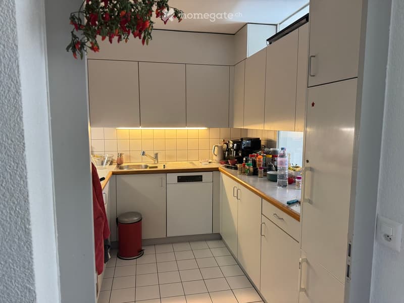 Compact kitchen with white cabinets, tiled floor, and various appliances including a sink, dishwasher, coffee maker, and refrigerator. Countertops hold several items such as bottles, cups, and a trash bin.