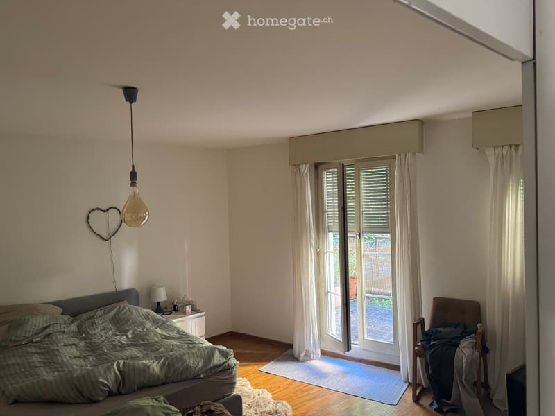 Cozy bedroom with wooden flooring, a large bed with green bedding, a nightstand with a lamp, a chair with folded clothes, and a heart-shaped wall decoration. Large sliding glass doors lead to a balcony with outdoor space visible.