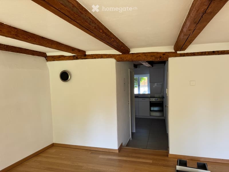 Empty room with wooden floor, white walls, exposed wooden beams on the ceiling, and a doorway leading to a modern kitchen area with grey cabinets and stainless steel appliances.