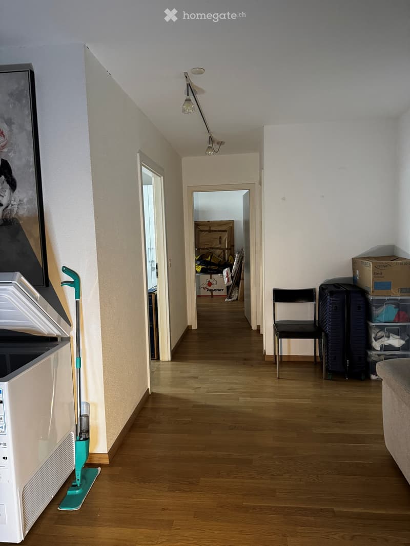 A narrow hallway with wooden flooring, white walls, and a slanted ceiling. A cleaning spray bottle is placed on the floor near the wall, and a black chair is positioned against the wall. A large mirror is mounted on the left wall, and several open doors l
