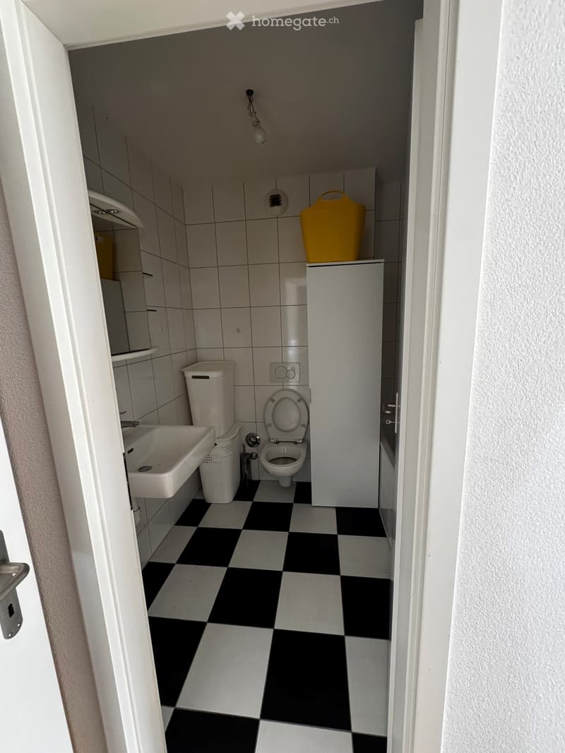 Small bathroom with black and white checkered floor, white tiled walls, toilet, sink, mirror, and a small cabinet with a yellow container on top.