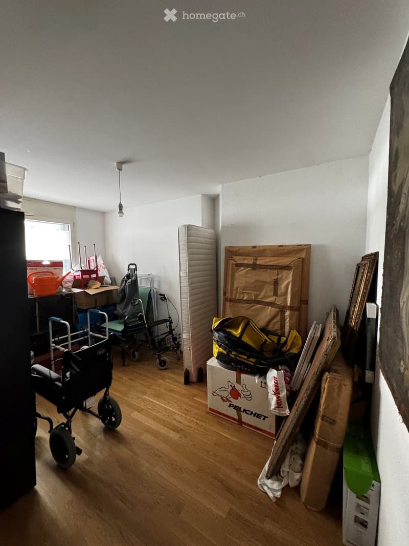 A room with wooden flooring, a baby stroller, a chair, a table, a backpack, a suitcase, and a futon.