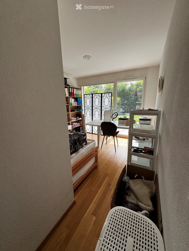 A furnished room with a wooden floor, a bed, a chair, a table, shelves, and a cat with its basket.