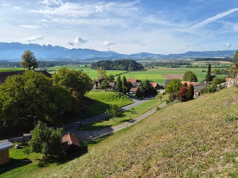 A panoramic view of a rural landscape with scattered houses, green fields, and distant mountains under a clear blue sky.