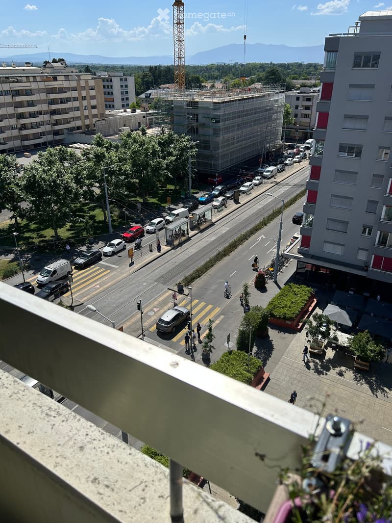 View from a balcony overlooking a busy urban street with cars, pedestrians, and cyclists. The scene includes modern buildings, a construction crane, and a clear view of the surrounding area.