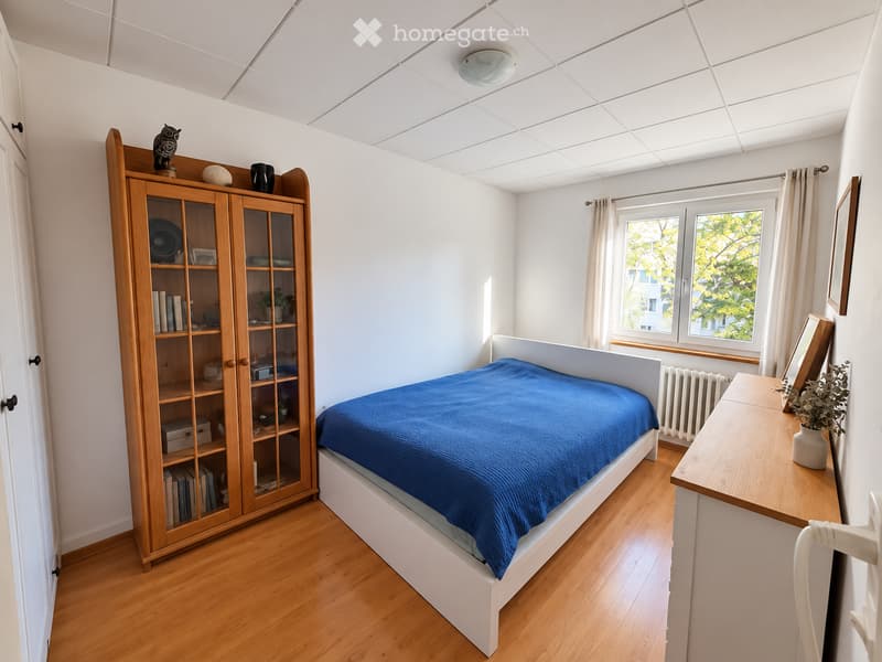 Bright bedroom with wooden floor, white walls, and large windows. Features a blue bedspread, wooden bookshelf, small table with a vase, and a mirror on the wall.