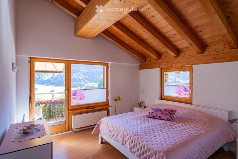 Cozy bedroom with wooden ceiling beams, large windows offering a scenic mountain view, a neatly made bed with pink bedding, a nightstand with a lamp, and a small table with decorative items.