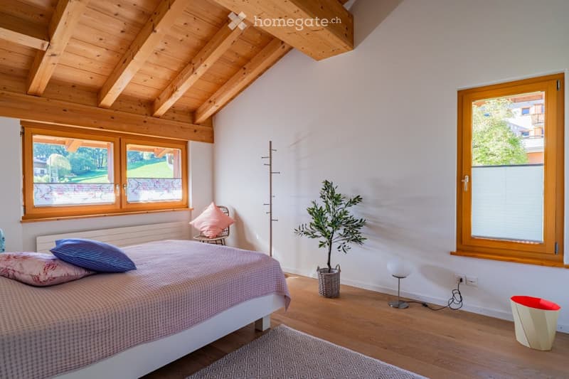 Cozy bedroom with wooden ceiling beams, large windows, a bed with pillows and a blanket, a chair with a cushion, a potted plant, and a floor lamp.
