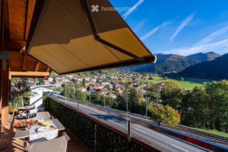Balcony with a scenic view of a town and mountains, featuring a small table with chairs, a bowl of fruit, and a cup.