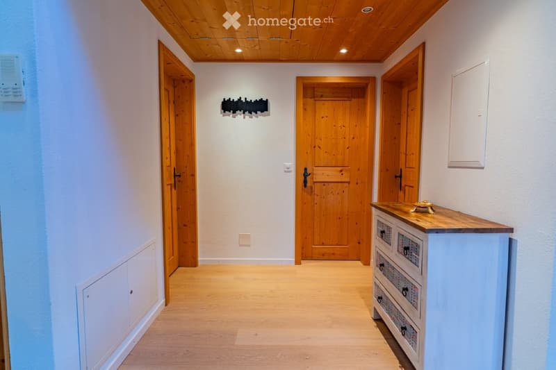A clean, well-lit hallway with wooden flooring, white walls, and several wooden doors leading to different rooms. A small console table with drawers is positioned on the right side, and a modern light fixture is mounted on the wall.