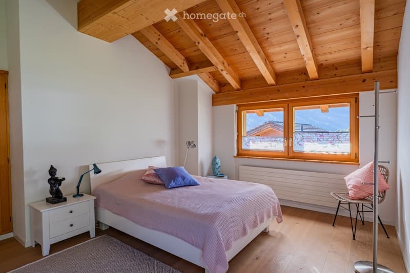 Cozy bedroom with wooden ceiling beams, large window, bed with pink and blue bedding, nightstand with lamp, decorative items, and a wicker chair.