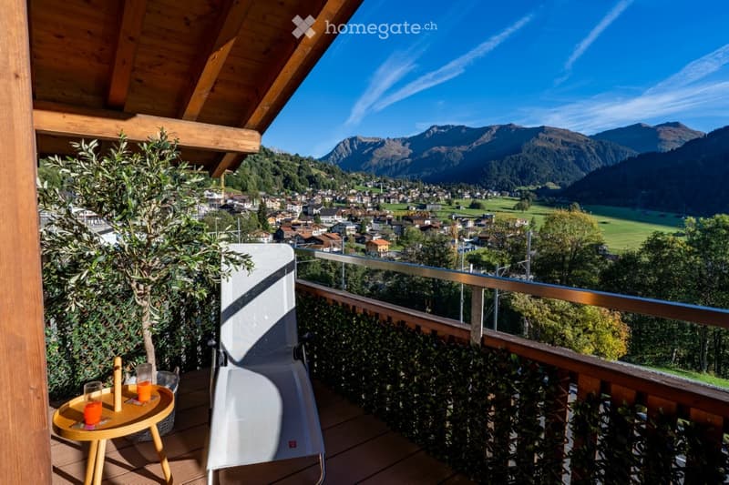 Balcony with wooden decking, small round table with drinks, white lounge chair, potted plants, and scenic mountain view of a town.