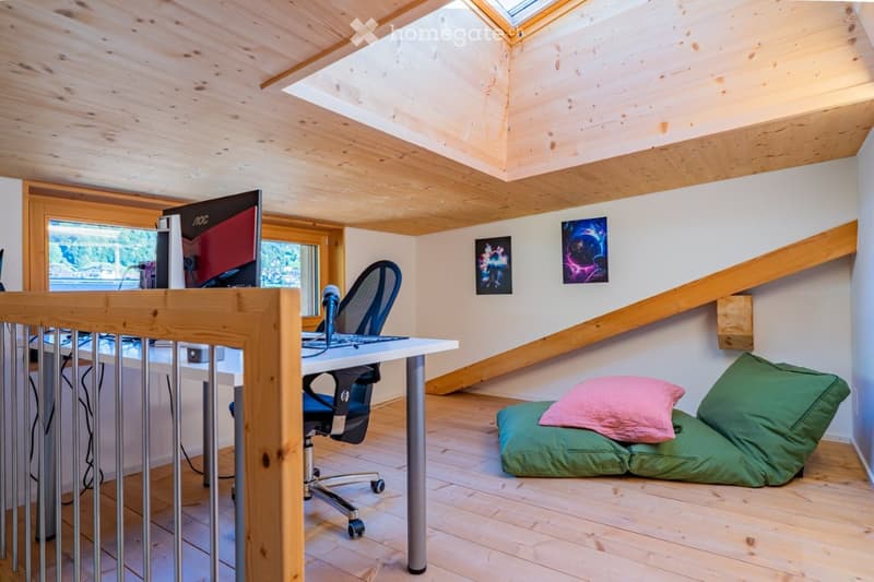 A cozy attic room with wooden beams and skylights, featuring a desk with a computer, a comfortable chair, and a small sofa with pillows.