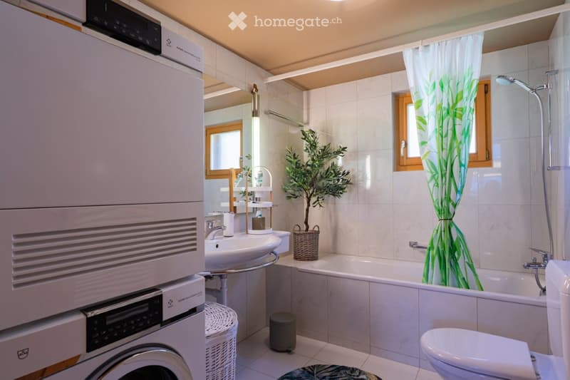 Modern bathroom with white tiles, a sink, bathtub, toilet, shower curtain, washing machine, and decorative plant in a wicker basket.