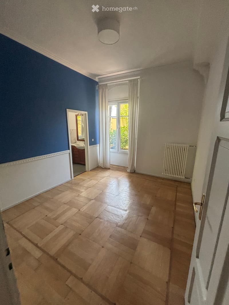 Empty room with wooden flooring, blue and white walls, a radiator, and a doorway leading to a bathroom.