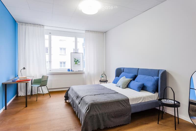 Modern bedroom with a large bed, nightstand, desk, and chair. Features a large window with white curtains, a blue accent wall, and a wooden floor.