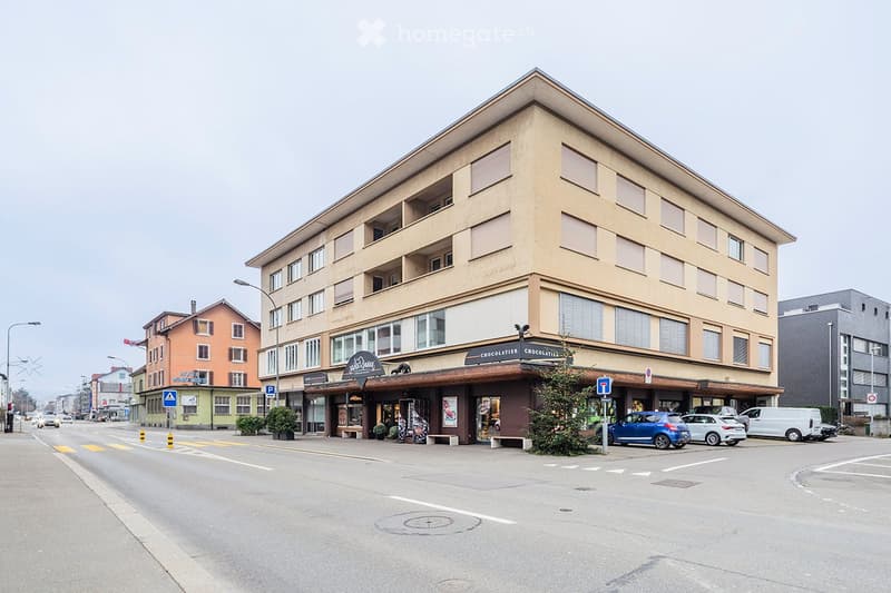 Multi-story building with a chocolate shop on the ground floor, several parked cars, and a street view.