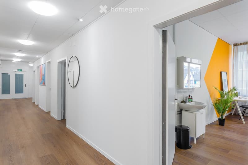 Modern hallway with white walls, wooden flooring, and recessed lighting. Features a round wall mirror, framed artwork, and a small adjacent bathroom with a sink, cabinet, and potted plant.
