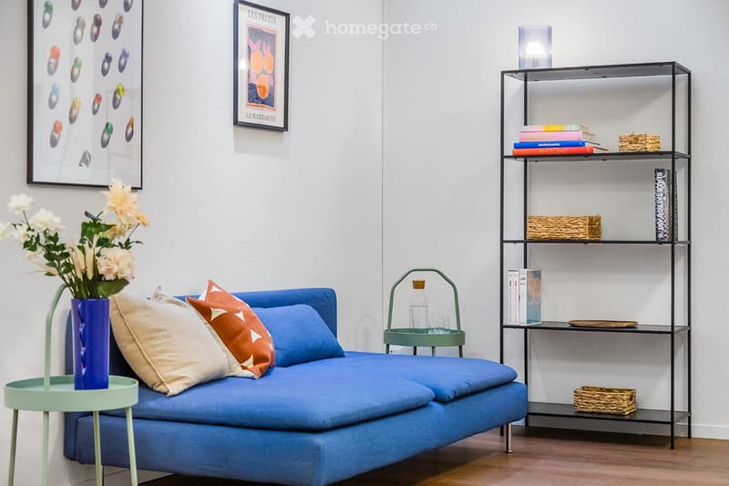 A modern living room with a blue sofa, decorative pillows, a small side table with a vase of white flowers, a metal shelf with books and decorative items, and framed artwork on the wall.