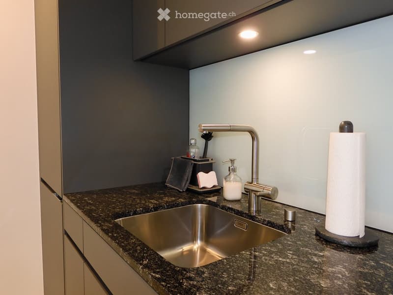 Modern kitchen sink area with granite countertop, stainless steel sink, sleek faucet, and various small items including a paper towel roll, soap dispenser, and a toothbrush holder.