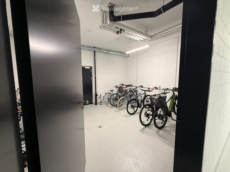 A clean, modern storage area with several bicycles lined up against the wall, featuring white walls, a concrete floor, and exposed pipes on the ceiling.