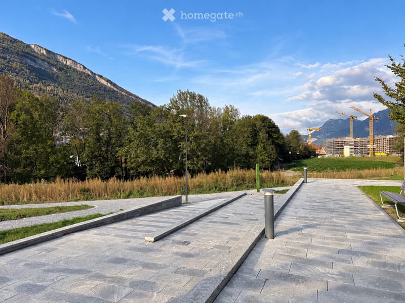 A paved outdoor area with stone tiles, low stone walls, and a bench, surrounded by trees and a mountain backdrop.