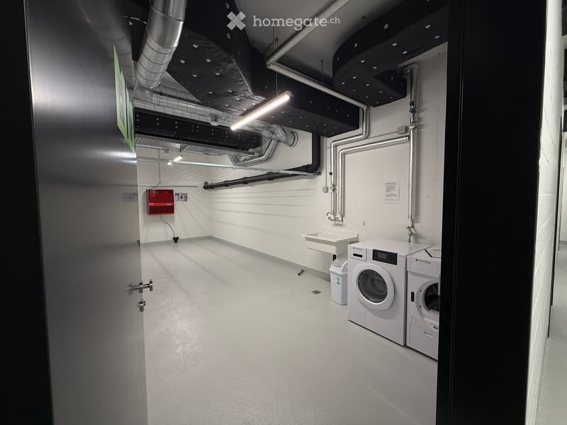 Laundry room with white washing machine, dryer, sink, and trash can. Exposed pipes and ductwork on the ceiling.