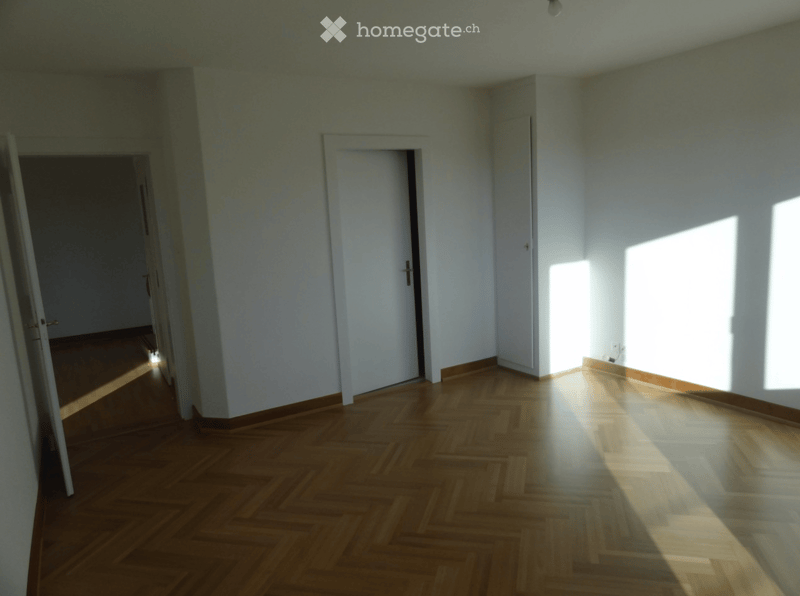Empty room with wooden parquet flooring, white walls, and natural light coming through a window on the right side. Two doors are visible, one leading to another room on the left and another closed door in the center.