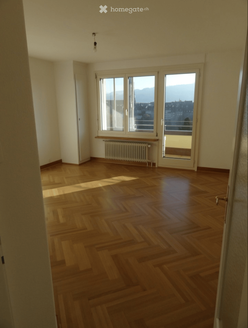 Empty room with wooden parquet flooring, large windows, radiator, and a view of the city and mountains.