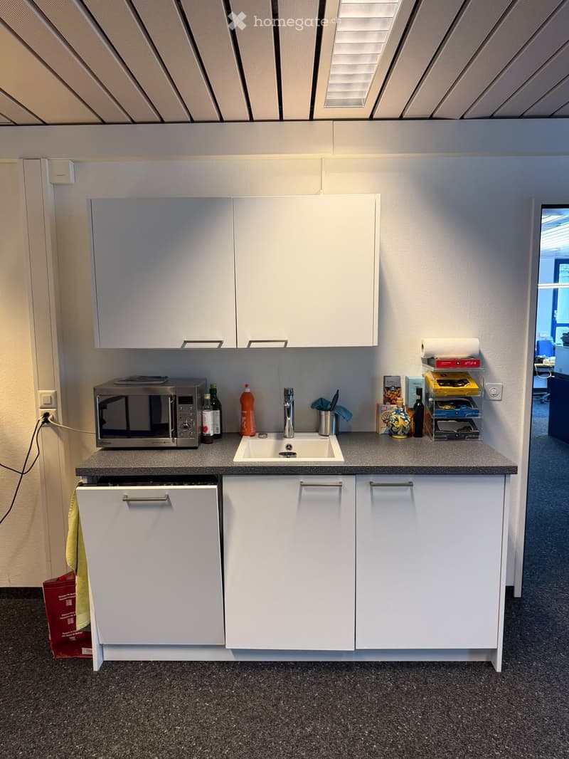 Compact kitchen with white cabinets, microwave, sink, dishwasher, and various small items on the counter.