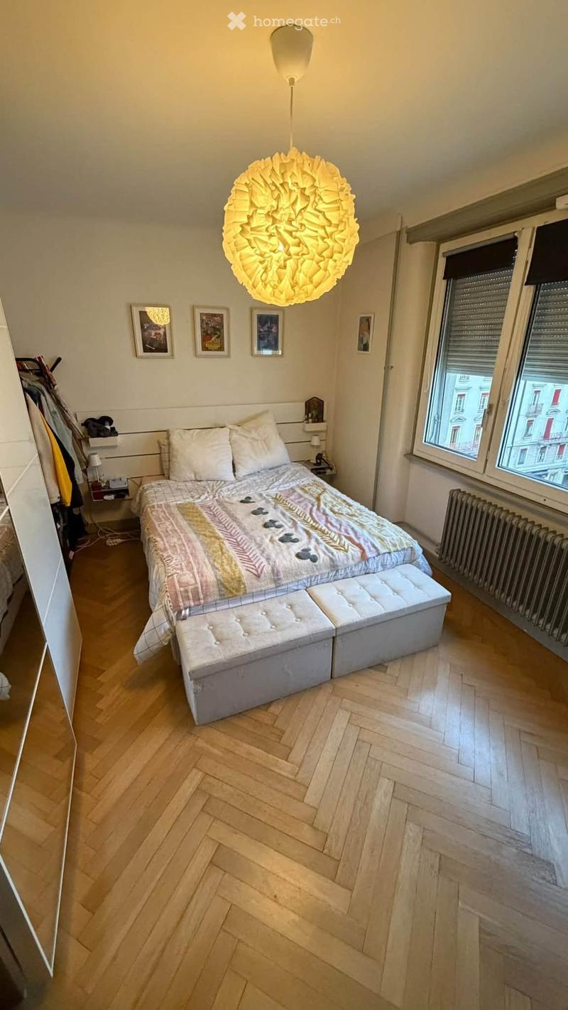 Cozy bedroom with wooden herringbone flooring, a large bed with patterned bedding, a matching bench at the foot, a nightstand with a lamp, and several picture frames on the wall. The room has large windows with blinds and a modern pendant light fixture.