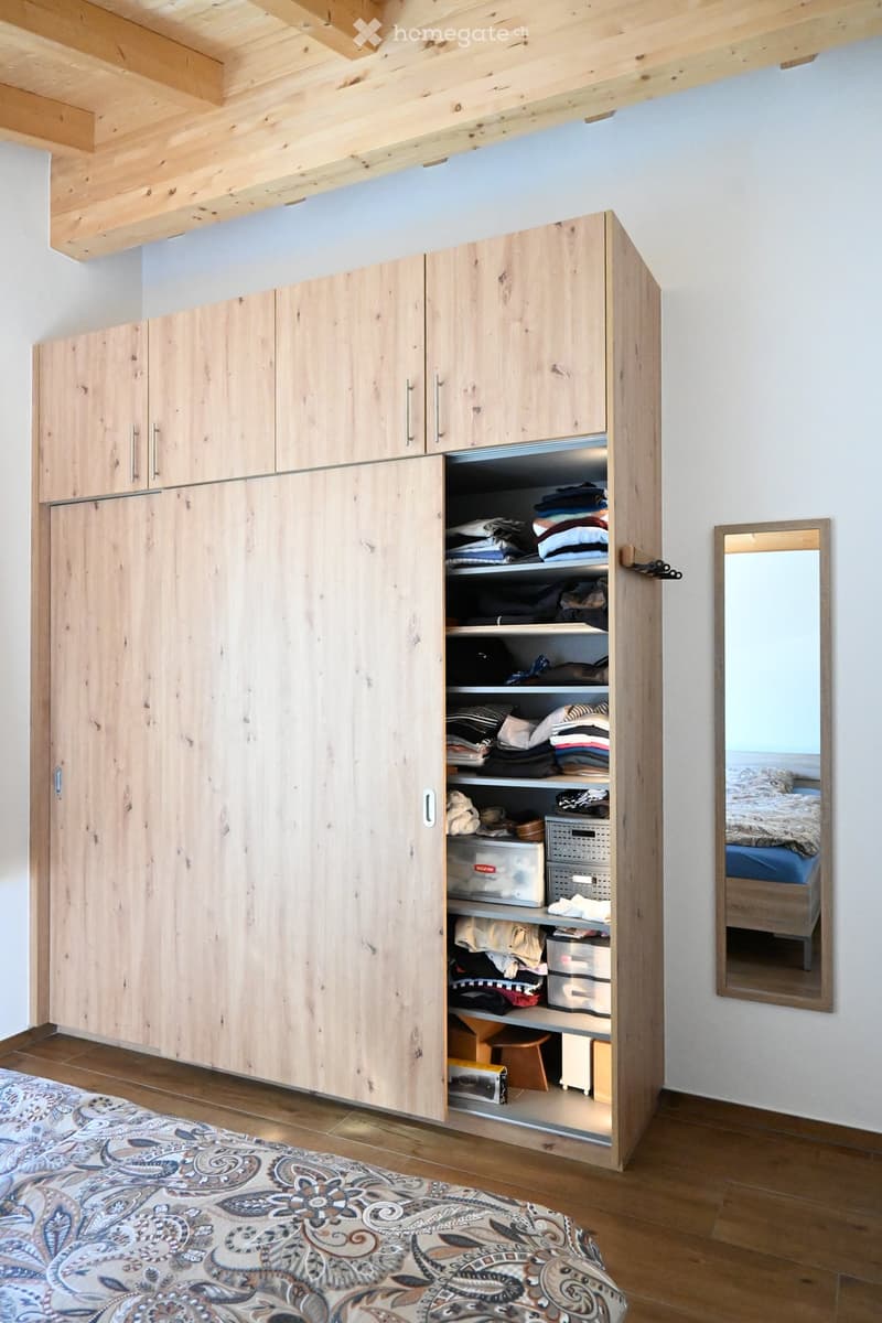 A modern bedroom with a large wooden wardrobe, a mirror, a bed with patterned bedding, and a decorative rug on the floor.