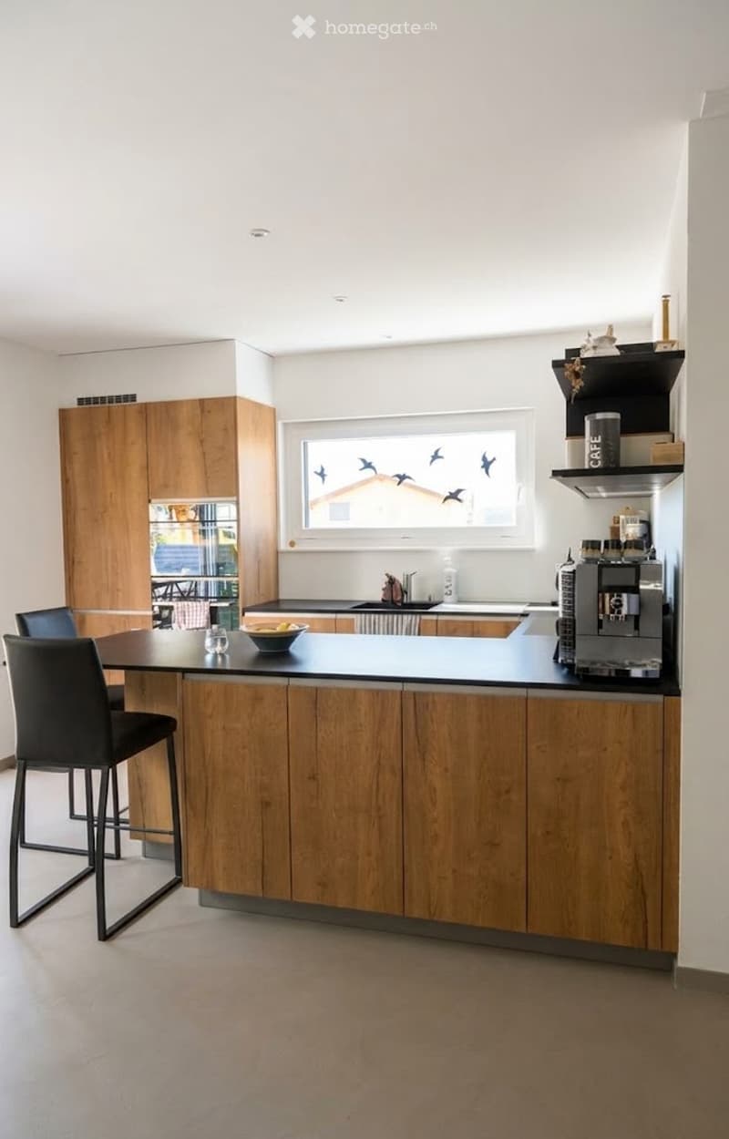 Modern kitchen with wooden cabinets, black island, two stools, coffee machine, and a window with birds painted on it.
