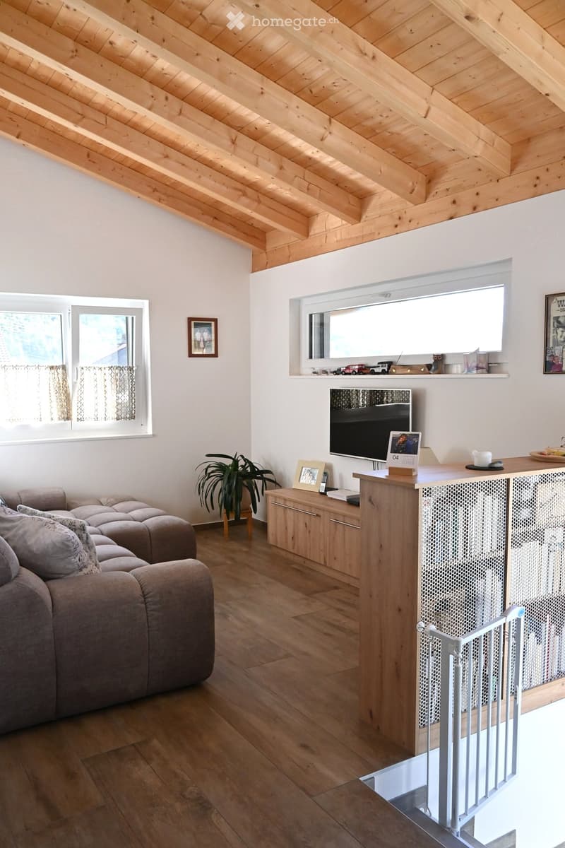 Bright living room with wooden ceiling, large windows, grey sectional sofa, coffee table, plant, TV, and decorative items on shelves.