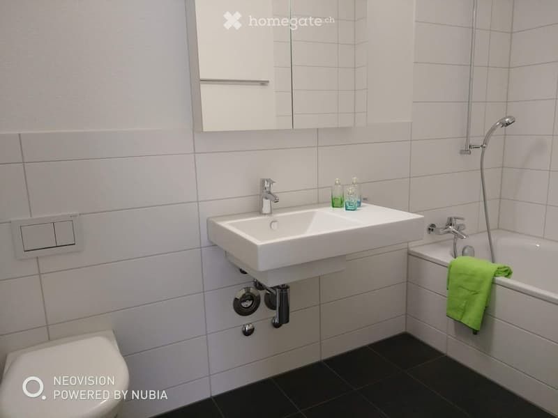 Modern bathroom with white tiles, a double sink, a bathtub, a toilet, a mirror, and a green towel on the side of the tub.