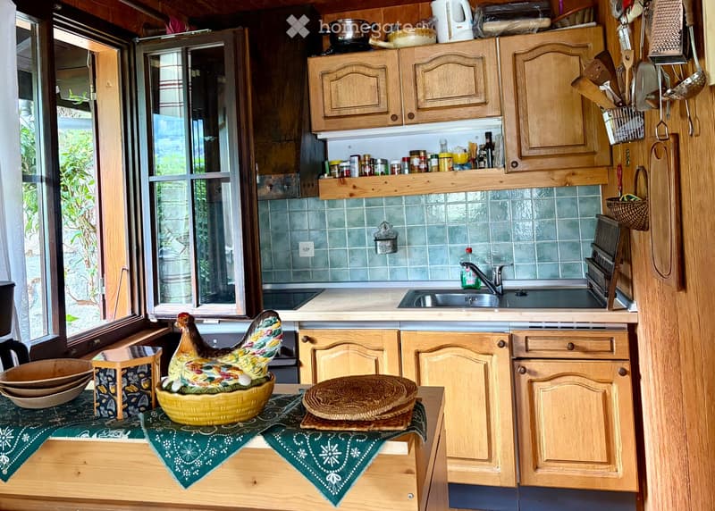 The kitchen features wooden cabinets, a sink with a faucet, and various kitchen utensils and appliances on the countertops. There is a decorative rooster figurine on a small table covered with a green cloth.