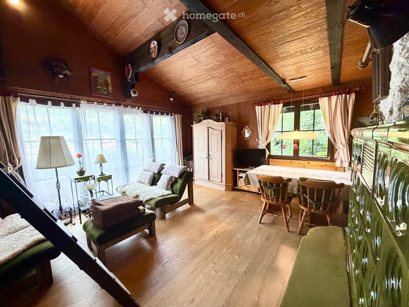 Cozy living room with wooden floors, exposed wooden beams, large windows with curtains, a green sofa, wooden chairs, a dining table, a television, and decorative items.
