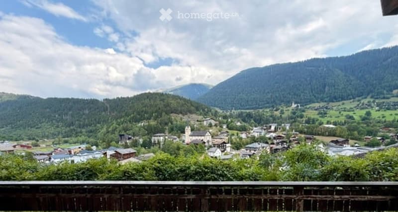 Panoramic view of a picturesque village nestled in a lush green valley surrounded by mountains, with scattered houses and a church visible.