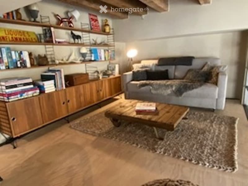 Modern living room with a grey sofa, wooden coffee table, bookshelf, and floor lamp. The room features exposed wooden beams on the ceiling and a cozy rug on the floor.