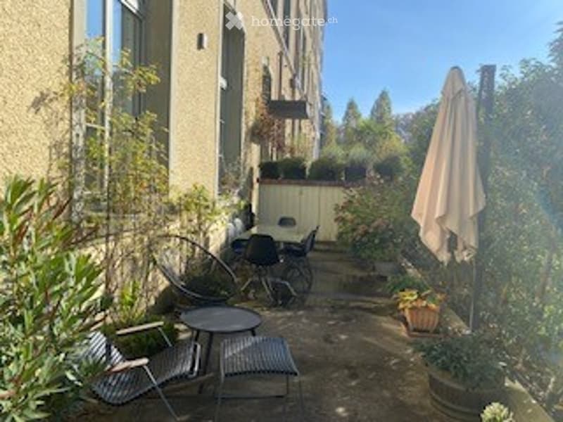 A small, cozy outdoor patio area with a table, chairs, and potted plants. The space is surrounded by greenery and features a closed umbrella leaning against a wall.