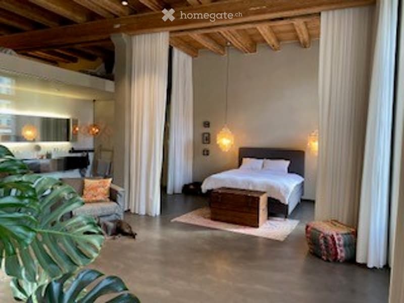 Open-concept living and bedroom area with exposed wooden beams, large windows, modern furniture including a sofa, ottoman, bed, nightstand, and a small table.