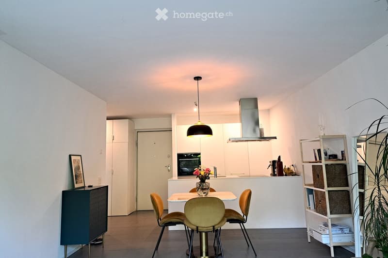 Open-plan living and dining area with modern furniture, including a dining table with four chairs, a cabinet, a shelf, and a vase with flowers on the table. The kitchen area features white cabinets, an oven, and a range hood.
