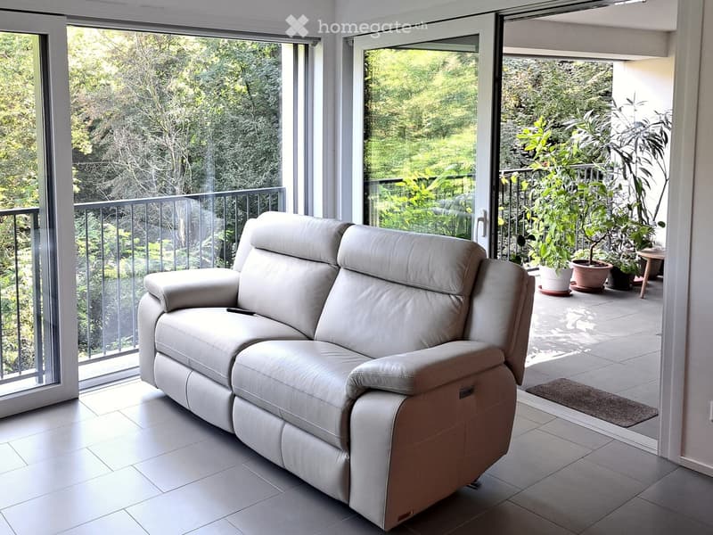 Modern living room with a white leather sofa, large windows, and a view of a lush garden through a balcony door.