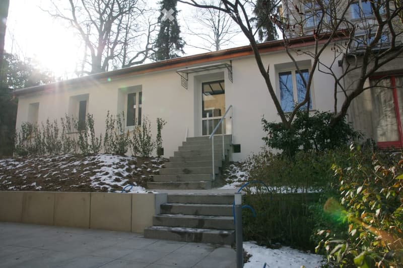 Single-story house with white walls, red roof, and a raised entrance accessed by concrete steps. The house has multiple windows and is surrounded by small shrubs and trees.