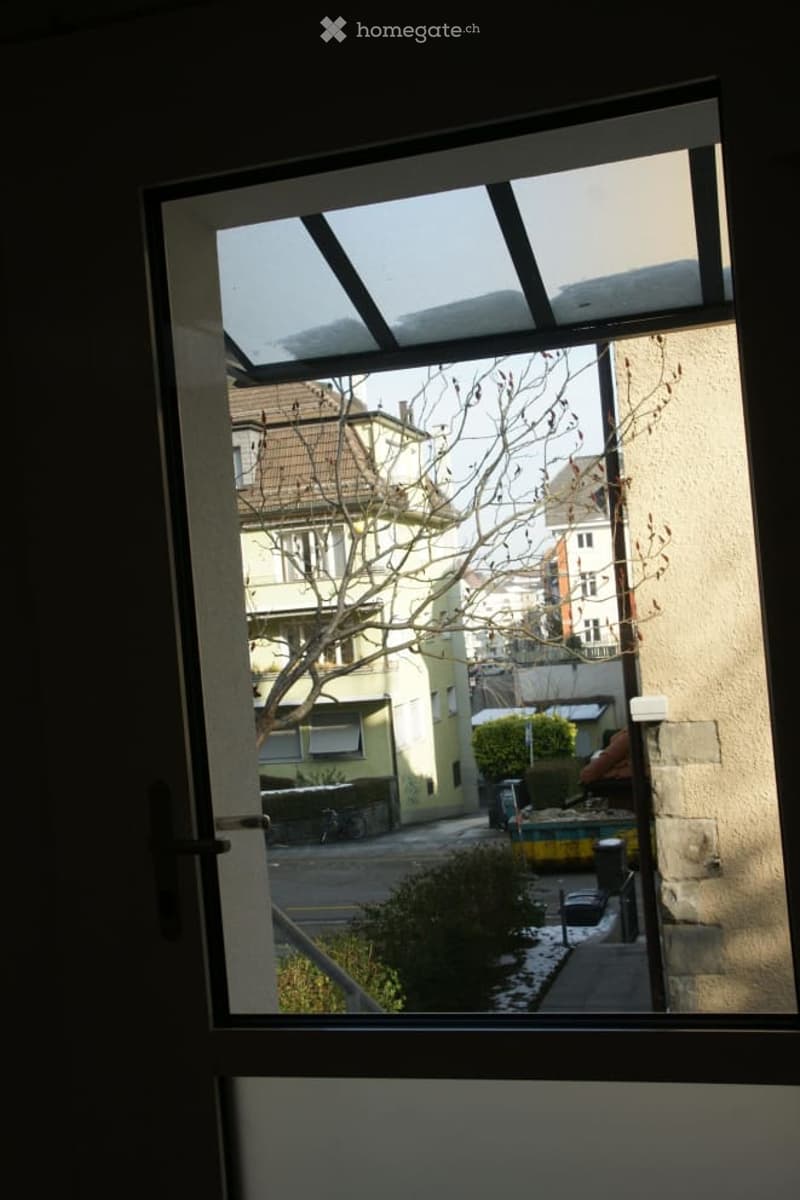 View from a balcony showing a street scene with buildings, trees, and parked cars.