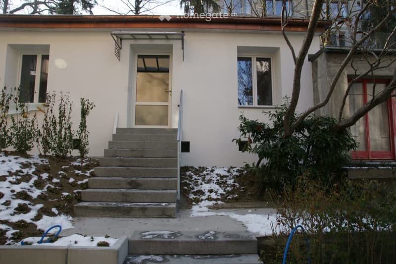 Two-story house with white exterior, raised ground floor, concrete steps leading to entrance, small garden area with snow patches.