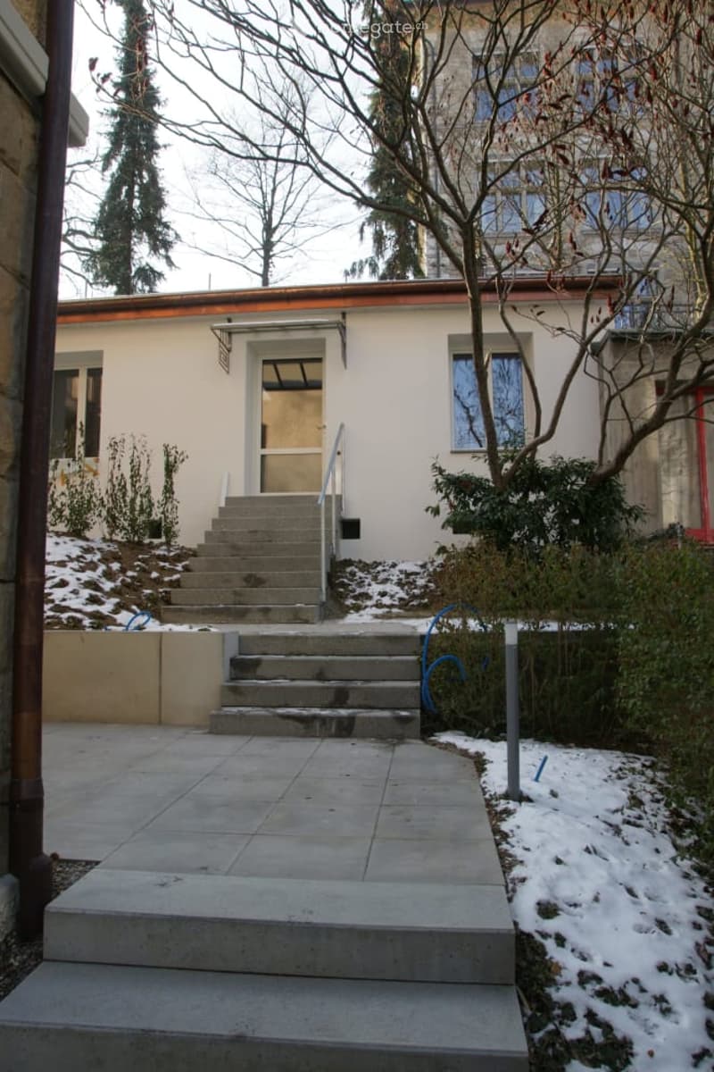 Two-story house with a red-tiled roof, white walls, and a concrete staircase leading to the entrance. The surrounding area includes snow-covered ground and some greenery.