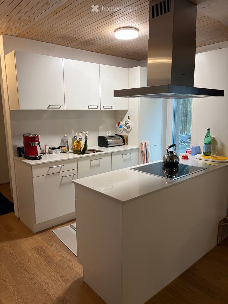 Modern kitchen with white cabinets, wooden flooring, and a central island with an induction stove. Features include a coffee maker, toaster, teapot, and various kitchen items on the countertops.