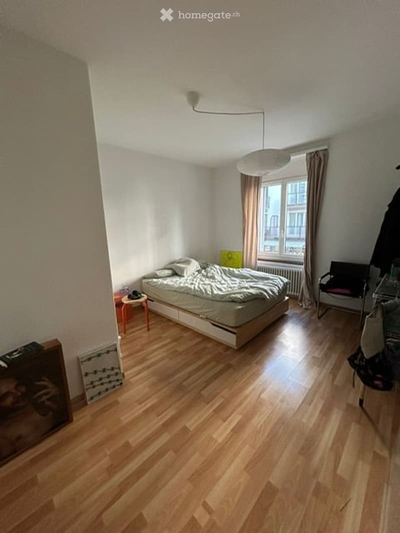 Cozy bedroom with wooden flooring, a double bed with pillows and blankets, a small nightstand, a chair, and a radiator near the window.